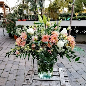 Es un centro de flor en florero de cristal, con margaritas dobles en tonos salmón, lisianthus blancos y color salmón, rosas blancas, y paniculata.